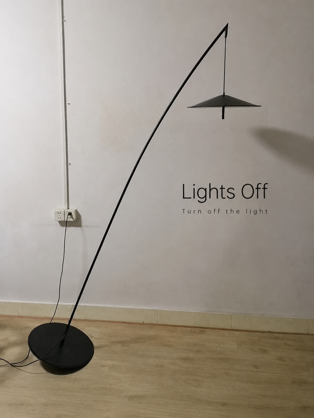 Black floor lamp against a white wall with text 'Lights Off' on a wooden floor.